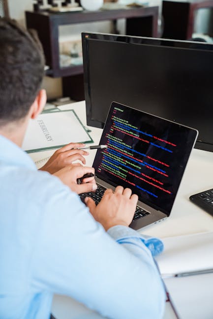 Close-up of a person's right hand using a laptop computer on a wooden desk. The hand is positioned over the keyboard, with fingers pressing keys. The laptop screen displays lines of coding or programm